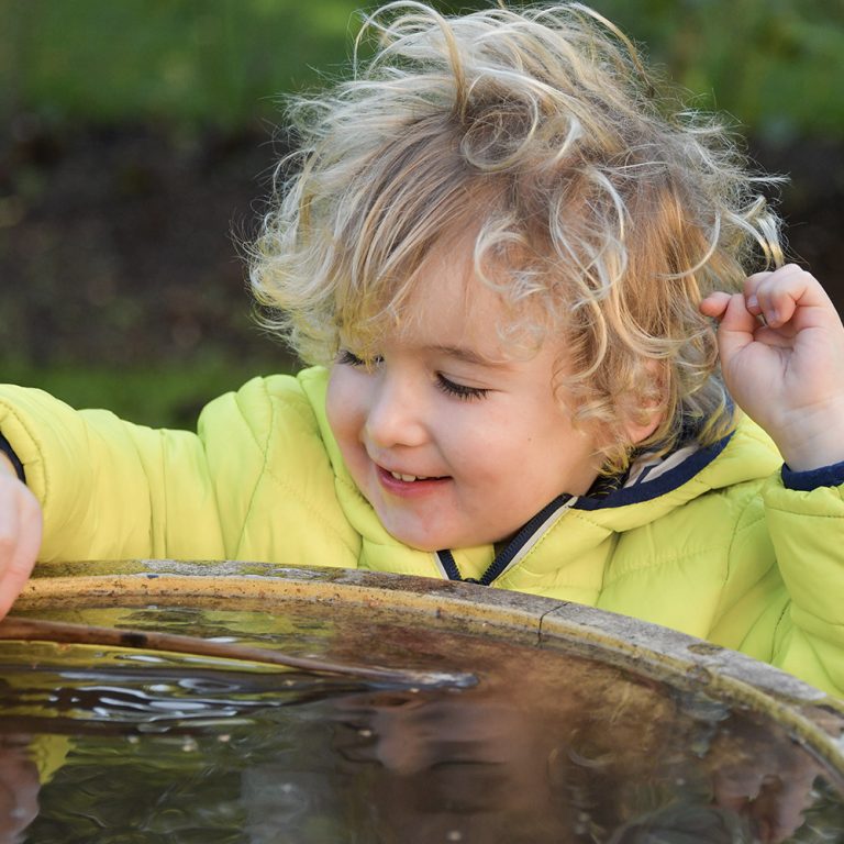 child wearing yellow coat putting a large leaf in the bird bath